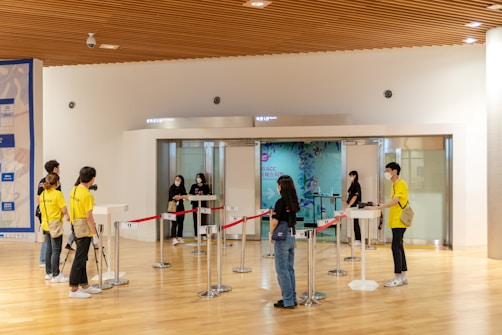 A group of people are gathered in a large hallway with a wooden ceiling. Some individuals are wearing yellow shirts labeled 'staff' and 'volunteer,' while others are dressed in casual clothing. Several are wearing face masks, suggesting a public setting. There are red ropes set up for line management and a digital check-in point at the entrance to a room. A large mural or poster can be seen in the background.