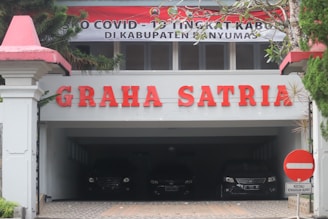 The image features the entrance of a building with the name 'GRAHA SATRIA' prominently displayed in red letters. Above the entrance, there's a banner with COVID-19 related messaging. Below the sign, several cars are parked in a shaded area. The surrounding area includes greenery and a traffic sign prohibiting entry, except for particular vehicles.