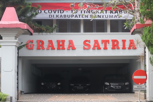 The image features the entrance of a building with the name 'GRAHA SATRIA' prominently displayed in red letters. Above the entrance, there's a banner with COVID-19 related messaging. Below the sign, several cars are parked in a shaded area. The surrounding area includes greenery and a traffic sign prohibiting entry, except for particular vehicles.