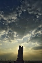 A silhouetted statue of an angel stands prominently against a dramatic sky filled with clouds. Rays of sunlight break through the clouds, casting an ethereal glow around the figure. The scene creates a striking contrast between the dark silhouette and the bright sky.