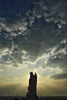 A silhouetted statue of an angel stands prominently against a dramatic sky filled with clouds. Rays of sunlight break through the clouds, casting an ethereal glow around the figure. The scene creates a striking contrast between the dark silhouette and the bright sky.