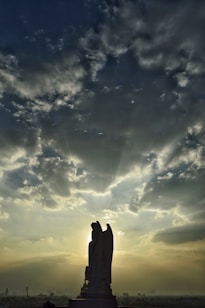 A silhouetted statue of an angel stands prominently against a dramatic sky filled with clouds. Rays of sunlight break through the clouds, casting an ethereal glow around the figure. The scene creates a striking contrast between the dark silhouette and the bright sky.