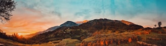 A scenic landscape featuring a lush, green valley with rolling hills and majestic mountains in the background. The sky is a vibrant blend of orange and blue hues, suggesting a sunrise or sunset. Small patches of forest and some isolated trees are visible, enhancing the natural beauty.
