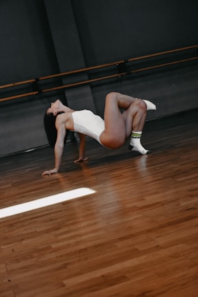 A person in a white leotard performs a graceful dance pose on a wooden floor in a studio with dark walls and a ballet barre. One leg is bent while the other extends with a foot wearing a white sock.