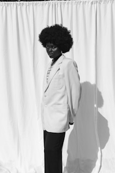 A person with a large, curly afro hairstyle is standing in front of a plain white curtain. They are wearing a light-colored blazer over a patterned shirt and dark pants. The lighting creates a sharp shadow on the curtain.