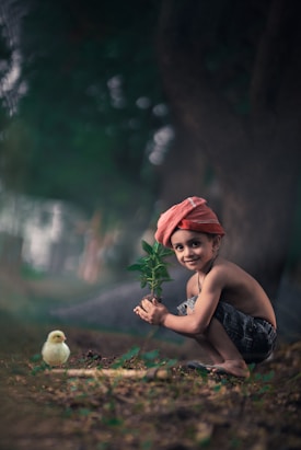 A young child crouches on the ground, holding a small plant with green leaves. The child is wearing a red headscarf and denim shorts, smiling towards the camera. A small yellow chick is near the child, surrounded by greenery and natural outdoor elements.