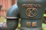 A close-up of a rusted, greenish-blue metal pipe with embossed markings, including the word 'TRADEMARK' and a circular symbol with a 'K' inside. Below, there are additional embossed characters in a different script.