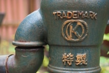 A close-up of a rusted, greenish-blue metal pipe with embossed markings, including the word 'TRADEMARK' and a circular symbol with a 'K' inside. Below, there are additional embossed characters in a different script.