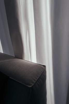 Soft, white curtains partially obscure the view, allowing light to filter through gently. A dark gray or black corner of a sofa or chair is visible in the foreground, aligning with the vertical lines of the drapery.