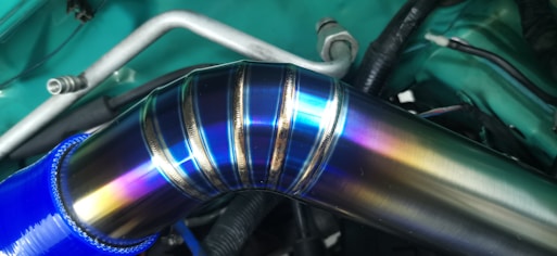 A metallic pipe with a colorful, iridescent surface that showcases shades of blue, purple, and yellow. The pipe is connected to a blue hose and is part of a vehicle's engine bay, featuring wires and metal components in the background.