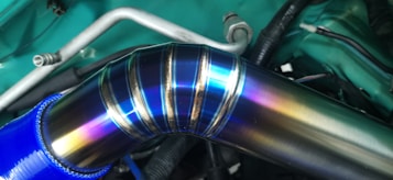 A metallic pipe with a colorful, iridescent surface that showcases shades of blue, purple, and yellow. The pipe is connected to a blue hose and is part of a vehicle's engine bay, featuring wires and metal components in the background.