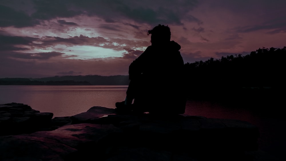日没時に水域の近くの岩の上に座っている男のシルエット