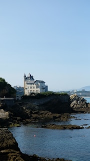 A picturesque coastal scene featuring a large, elegant mansion perched on rocky cliffs overlooking the sea. The building has a traditional architectural style with towers and multiple windows. The rocky shoreline is surrounded by calm waters, and the horizon shows distant mountains.