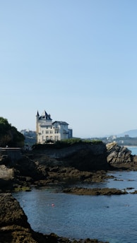 A picturesque coastal scene featuring a large, elegant mansion perched on rocky cliffs overlooking the sea. The building has a traditional architectural style with towers and multiple windows. The rocky shoreline is surrounded by calm waters, and the horizon shows distant mountains.
