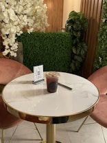 A round marble-top table with golden edges is placed in a cozy corner with two pink upholstered chairs. On the table, there is a plastic cup containing iced coffee, a black straw, and a holder displaying a QR code. The background features lush green plants, including a green wall and a faux cherry blossom tree, with wooden paneling.