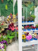 A vibrant display of various flowers in a shop setting with multiple shelves filled with bouquets in blue containers. The background features a lush green wall made of artificial foliage. The assortment includes roses, sunflowers, lilies, and other colorful flowers, creating a lively and lush atmosphere.