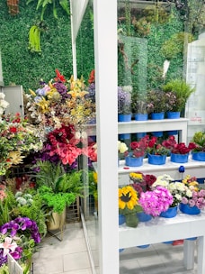 A vibrant display of various flowers in a shop setting with multiple shelves filled with bouquets in blue containers. The background features a lush green wall made of artificial foliage. The assortment includes roses, sunflowers, lilies, and other colorful flowers, creating a lively and lush atmosphere.