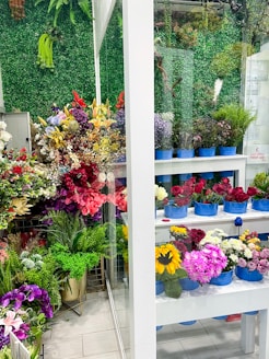 A vibrant display of various flowers in a shop setting with multiple shelves filled with bouquets in blue containers. The background features a lush green wall made of artificial foliage. The assortment includes roses, sunflowers, lilies, and other colorful flowers, creating a lively and lush atmosphere.