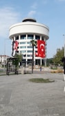 A round, modern building with multiple floors, featuring large Turkish flags and a portrait banner. The structure is adorned with white cladding and has large windows. A stone-paved open area with palm trees and benches is in the foreground.