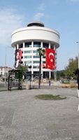 A round, modern building with multiple floors, featuring large Turkish flags and a portrait banner. The structure is adorned with white cladding and has large windows. A stone-paved open area with palm trees and benches is in the foreground.