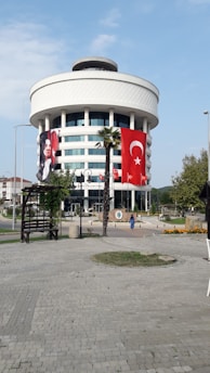 A round, modern building with multiple floors, featuring large Turkish flags and a portrait banner. The structure is adorned with white cladding and has large windows. A stone-paved open area with palm trees and benches is in the foreground.
