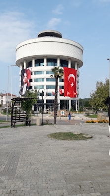 A round, modern building with multiple floors, featuring large Turkish flags and a portrait banner. The structure is adorned with white cladding and has large windows. A stone-paved open area with palm trees and benches is in the foreground.