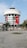 A round, modern building with multiple floors, featuring large Turkish flags and a portrait banner. The structure is adorned with white cladding and has large windows. A stone-paved open area with palm trees and benches is in the foreground.