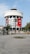 A round, modern building with multiple floors, featuring large Turkish flags and a portrait banner. The structure is adorned with white cladding and has large windows. A stone-paved open area with palm trees and benches is in the foreground.