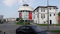 A modern cylindrical building with several floors is adorned with a large Turkish flag. Nearby, there is a traditional-looking building with black and white contrasts, appearing to be historical or of cultural significance. The scene includes a car parked on the street, and a clear sky provides a backdrop.