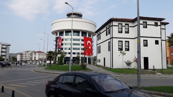 A modern cylindrical building with several floors is adorned with a large Turkish flag. Nearby, there is a traditional-looking building with black and white contrasts, appearing to be historical or of cultural significance. The scene includes a car parked on the street, and a clear sky provides a backdrop.