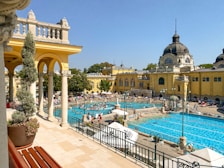 An expansive outdoor pool area surrounded by historic architecture with beige and yellow buildings featuring domed roofs. People enjoy the sun and swim in the pools, with umbrellas and loungers scattered around. The scene is vibrant with activity on a bright, clear day.