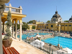 An expansive outdoor pool area surrounded by historic architecture with beige and yellow buildings featuring domed roofs. People enjoy the sun and swim in the pools, with umbrellas and loungers scattered around. The scene is vibrant with activity on a bright, clear day.