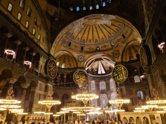 A grand interior with ornate chandeliers illuminating the space. The ceiling features intricate gold and blue designs and large circular calligraphic panels with Arabic inscriptions. The architecture is a blend of Byzantine and Islamic styles, reflecting a historical and cultural richness.