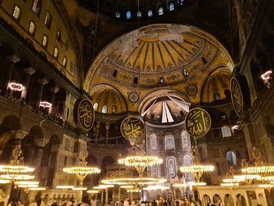 A grand interior with ornate chandeliers illuminating the space. The ceiling features intricate gold and blue designs and large circular calligraphic panels with Arabic inscriptions. The architecture is a blend of Byzantine and Islamic styles, reflecting a historical and cultural richness.