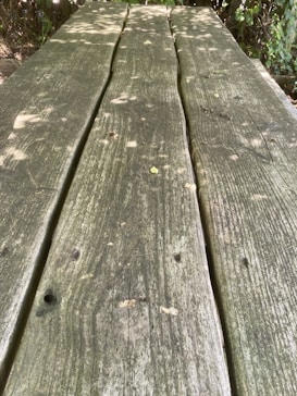 Weathered and sun-dappled wooden planks are aligned, forming a natural table surface. The texture of the wood is rough, with visible grain patterns and knots. Light filters through nearby foliage, casting scattered shadows and highlights.