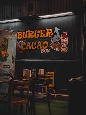 A dimly lit restaurant interior features a wall with bold yellow and orange lettering advertising 'Burger and Cacao Box.' The decor includes subtle illustrations of fries, a cup of hot chocolate, and a soda cup. A row of wooden chairs and tables are arranged in the foreground, creating a cozy and casual dining atmosphere.