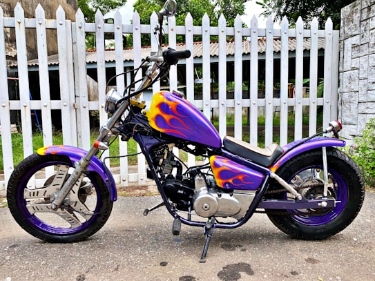 A custom motorcycle with a vibrant purple body featuring flame designs in yellow and orange. The bike is parked on a paved surface in front of a white picket fence. The handlebars and mechanical details are visible, and the wheels have matching purple rims.