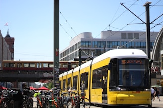 A yellow tram moves along a city street, with parked bicycles lined up along the sidewalk. In the background, there is an elevated railway track with a train passing by. Modern buildings and a few outdoor café umbrellas are also visible under a clear blue sky.