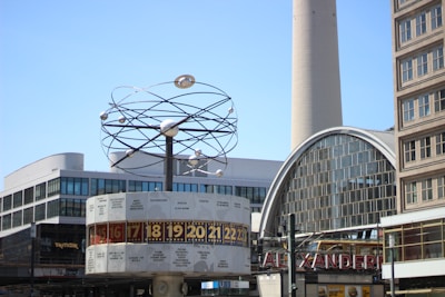 A large, outdoor clock displays different time zones beneath a structure resembling an astronomical sculpture. The surrounding area includes modern and historical buildings, one with a curved glass facade and a tall tower in the background.