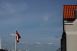 A Danish flag flutters in the breeze against a blue sky, with a few clouds scattered throughout. Wind turbines can be seen in the distant background. In the foreground, part of a building is visible, featuring a red-tiled roof.