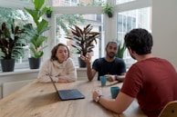Three people are seated around a wooden table having a discussion. There are several potted plants placed on the window sill behind them, letting in natural light through large windows. Two of the individuals hold blue mugs, and a laptop is open on the table. The room has a relaxed and collaborative atmosphere.