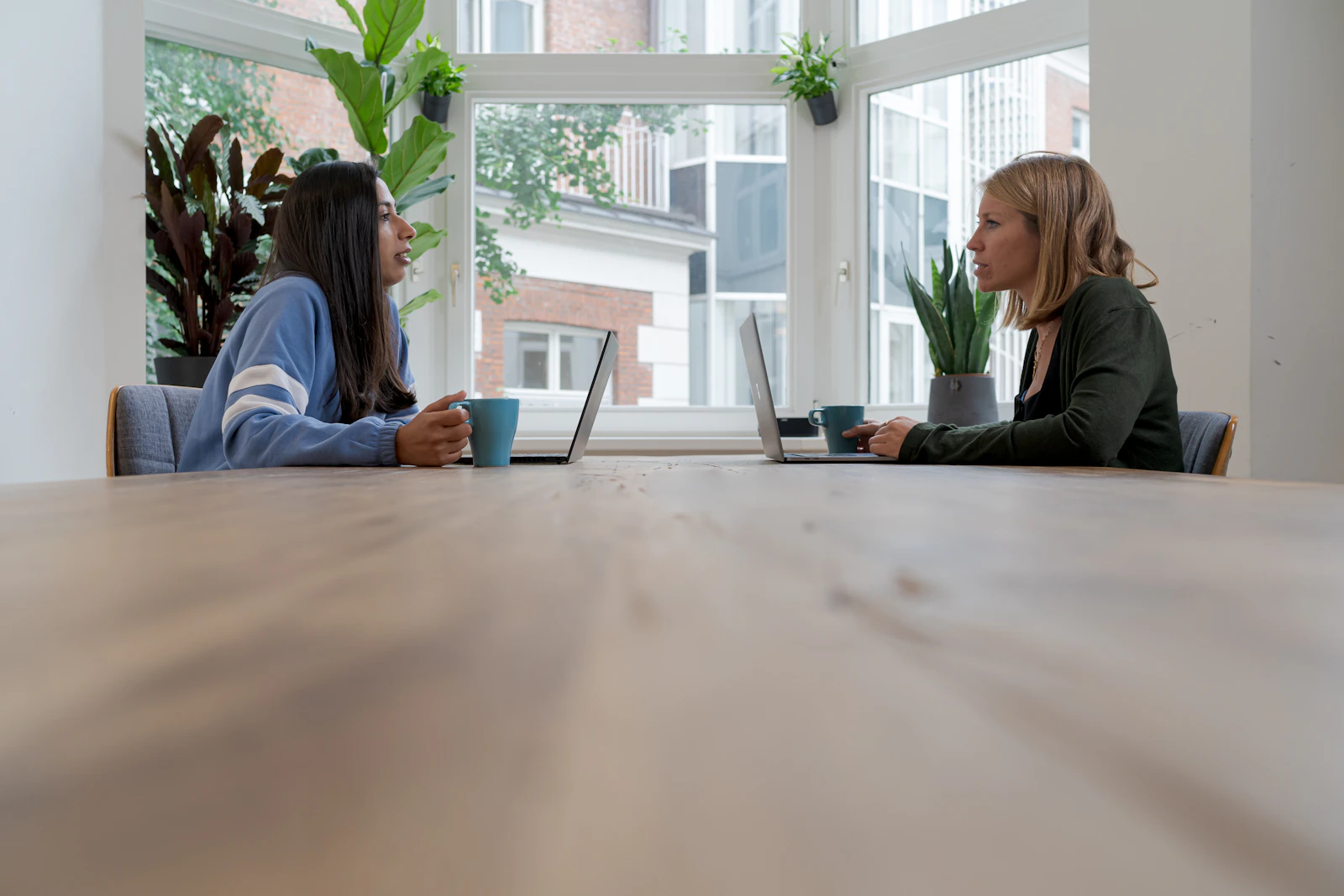 Two people talking at a long pale table by a window