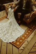 A couple sits together on a vintage floral-patterned sofa. The woman's white lace wedding dress with intricate floral designs trails elegantly onto a rustic wooden floor. A rich, patterned rug with tassels adds warmth to the setting. The man wears a dark suit and rests one hand on the armrest. The scene conveys a classic and cozy atmosphere.