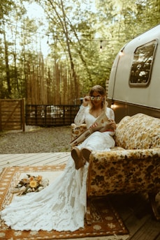 A woman in a lacy white dress lounges on a floral-patterned vintage couch outdoors. She holds a magazine and adjusts her sunglasses. The setting includes a rug with a bouquet of flowers on it, surrounded by a forest and a vintage trailer.