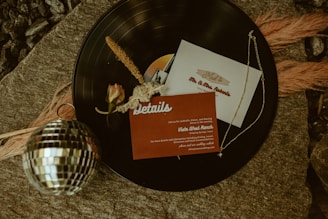 A vinyl record serves as a backdrop for several items, including an invitation card, a small rose, and a delicate gold chain. A disco ball is placed in the foreground, while feathery decorations and stones create a rustic and elegant scene.
