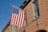 A brick building features an American flag mounted on a pole, with clear blue skies in the background. The building has multiple windows with white trim, and the facade consists of reddish-brown bricks.