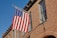 A brick building features an American flag mounted on a pole, with clear blue skies in the background. The building has multiple windows with white trim, and the facade consists of reddish-brown bricks.