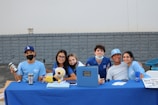 A group of six young individuals are sitting at a table covered with a blue tablecloth. They are smiling and appear to be part of a club or school event. Some are wearing blue shirts and hats with various logos. On the table, there are a few items such as a laptop, a stuffed toy, drink containers, and some papers.