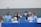 A group of six young individuals are sitting at a table covered with a blue tablecloth. They are smiling and appear to be part of a club or school event. Some are wearing blue shirts and hats with various logos. On the table, there are a few items such as a laptop, a stuffed toy, drink containers, and some papers.