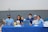 A group of six young individuals are sitting at a table covered with a blue tablecloth. They are smiling and appear to be part of a club or school event. Some are wearing blue shirts and hats with various logos. On the table, there are a few items such as a laptop, a stuffed toy, drink containers, and some papers.
